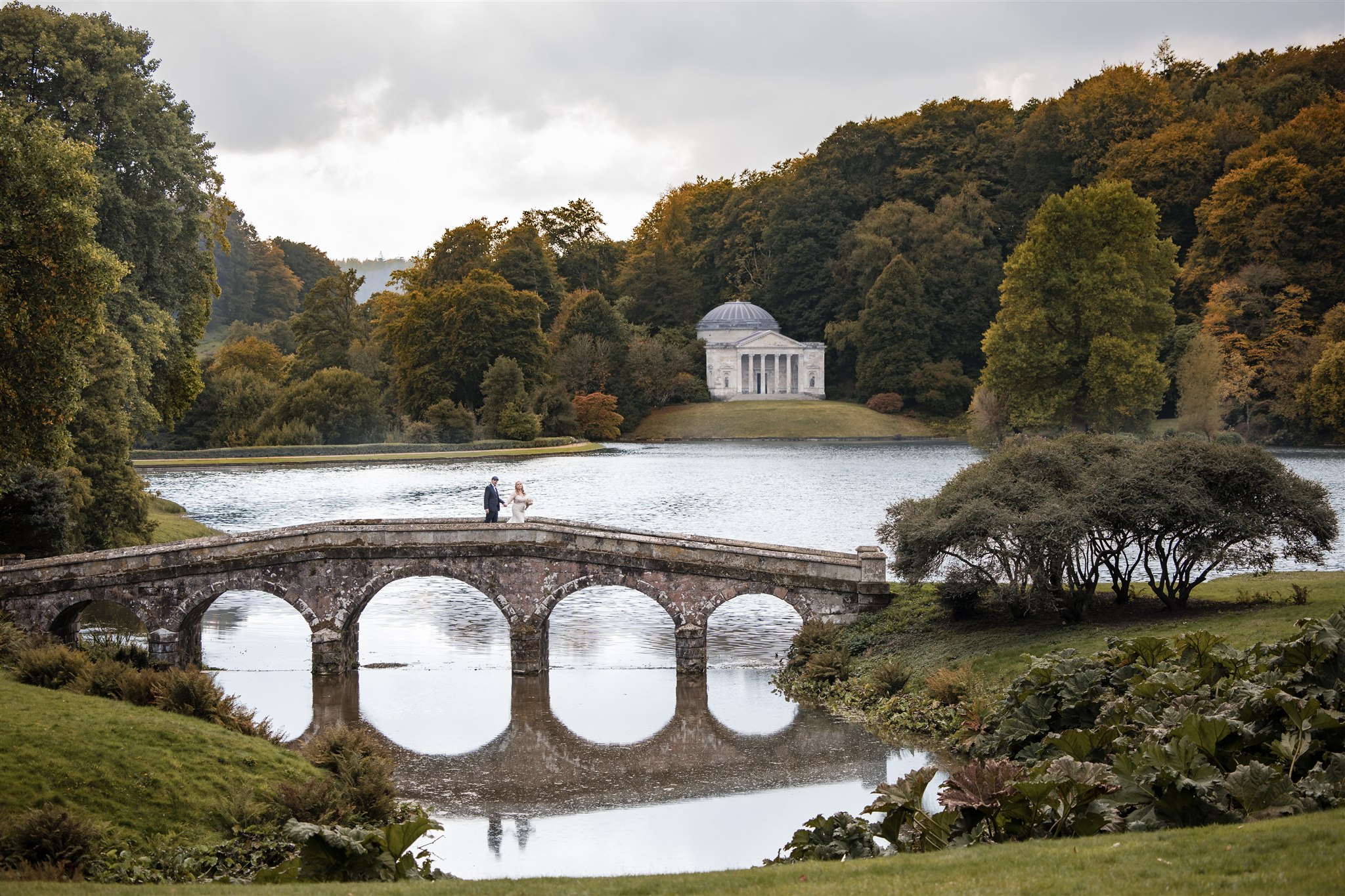 Saffron and Ryan walking across the bridge at Stourhead during their autumn wedding.