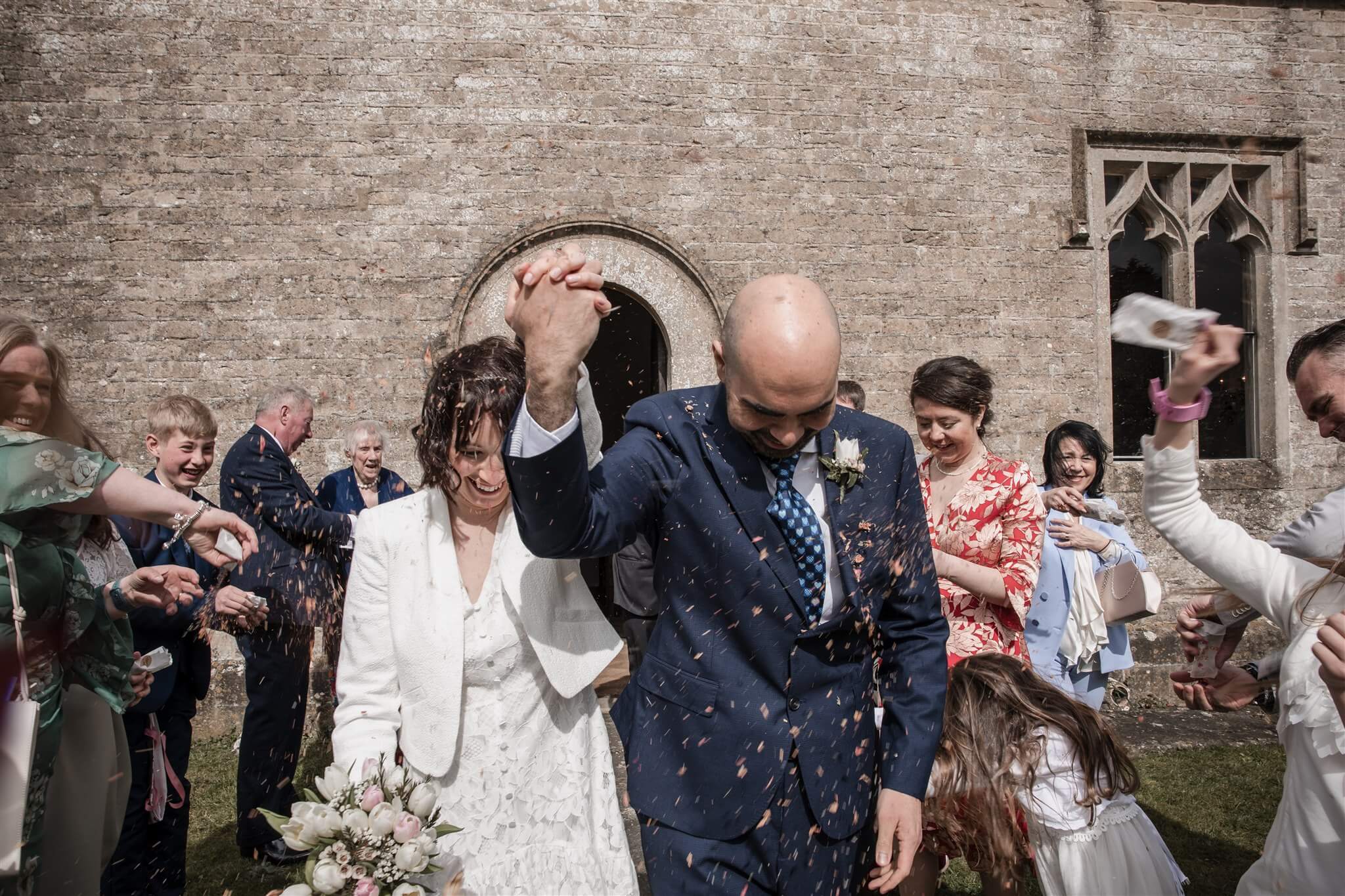 Zoe and Rodolfo walking through confetti outside St Osmund’s Chapel in Tarlton during their Cotswolds wedding.